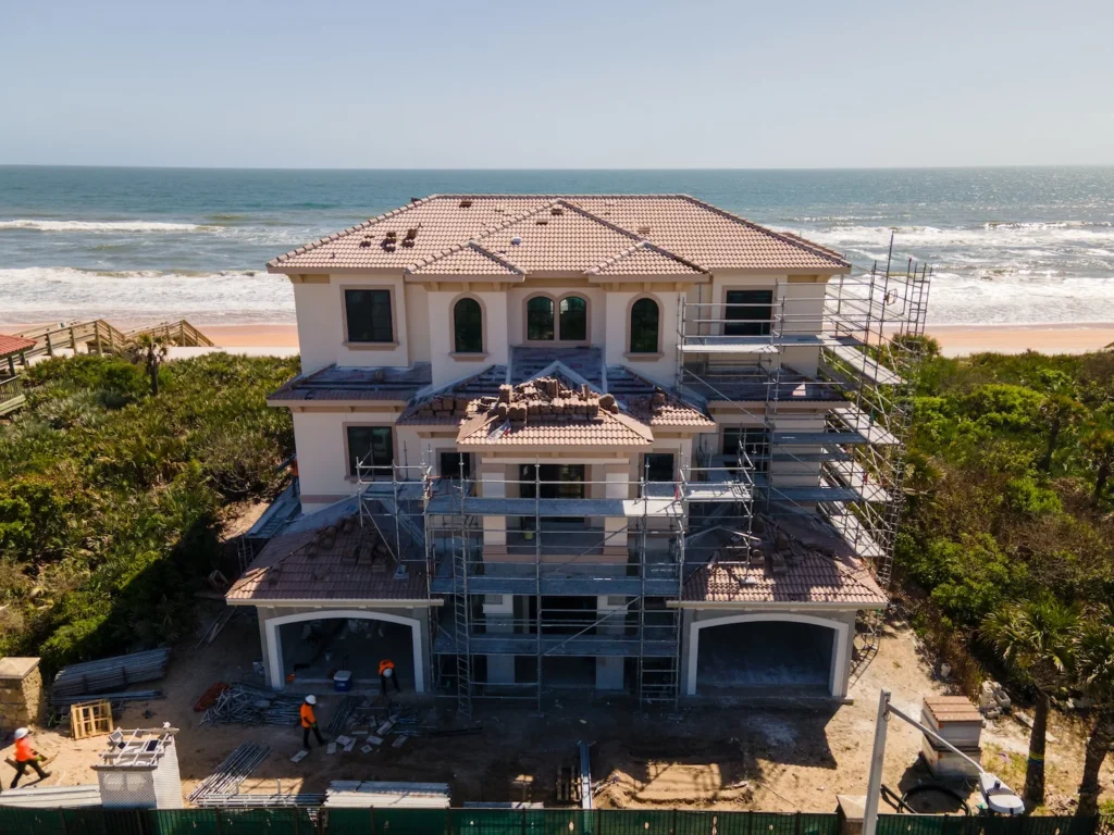 A new luxury home under construction in Palm Coast, FL seen from above - scaffolding is setup around the home and workers are entering the home. The home is situated on the beach by the water